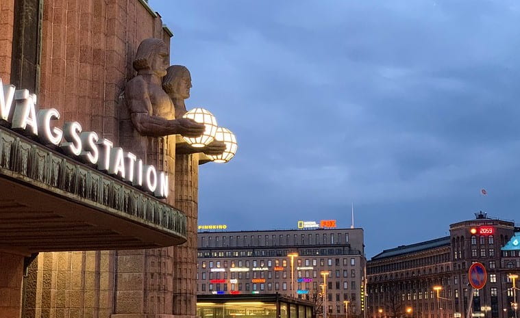 The main railway station in Helsinki, featuring art deco motifs, including tall streamlined human figurines holding lighting fixtures, toward dusk with other buildings in the lower right, with cloud cover overhead.