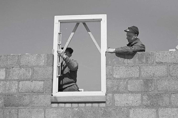 In a black and white photo, two men construct a window frame into a wall.